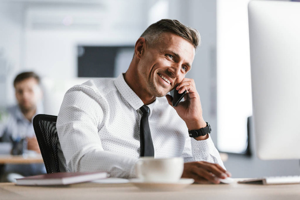 Image of european adult man 30s wearing white shirt and tie sitting at desk in office by computer and speaking on smartphone - Фото, изображение