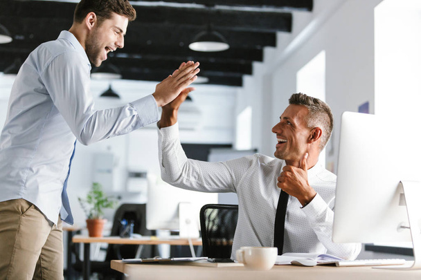 Photo of cheerful businessmen 30s in formal clothes giving high five together in office during successful deal - Foto, Imagen