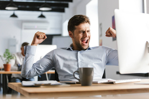 Joyful male manager rejoices and using computer while sitting by the table in office - Photo, Image