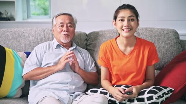 asian granddaughter with remote control and grandfather sitting on couch watching television indoors - Materiaali, video