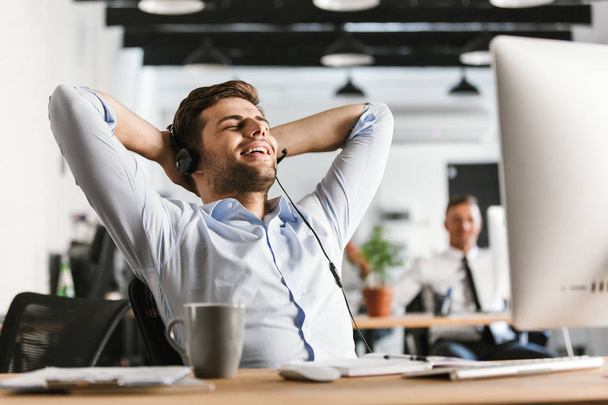 Joyful office manager talking by headset with microphone while sitting with closed eyes by the table in office - Foto, Imagem