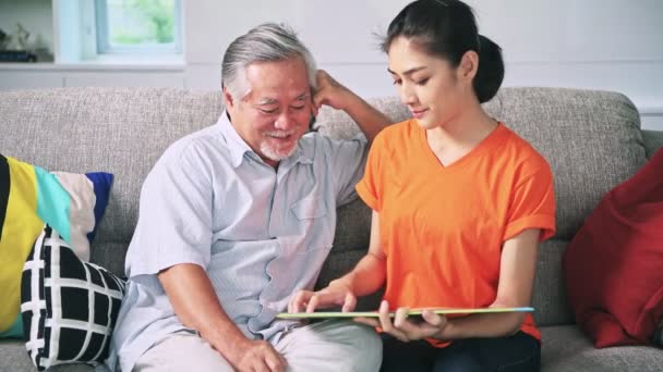asian grandfather with grey hair and granddaughter sitting on couch smiling talking and reading book  - Materiaali, video