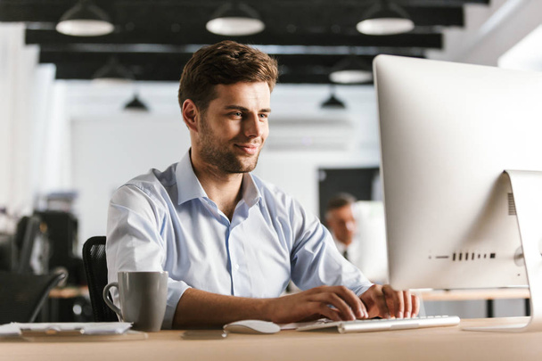Image of Smiling business man using computer while sitting by the table in office - Foto, Imagem