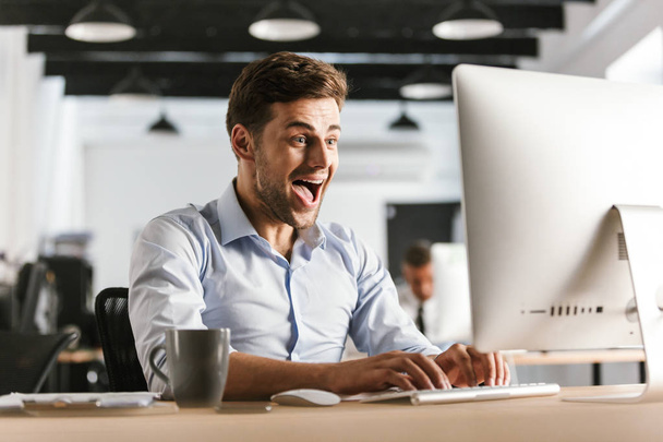 Happy screaming business man using computer and rejoices while sitting by the table in office - Foto, imagen