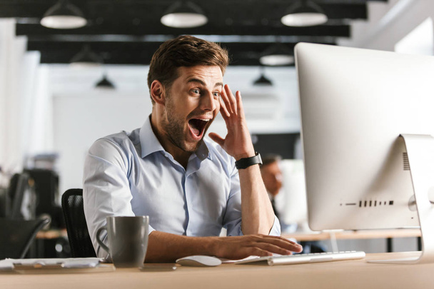 Surprised screaming business man using computer while sitting by the table in office - Fotó, kép