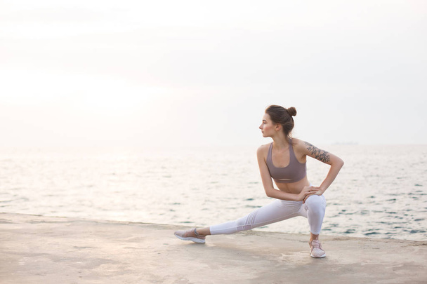 Menina atenciosa no topo desportivo e leggings brancos praticando ioga com incrível vista para o mar no fundo. Jovem mulher se alongando junto ao mar
 - Foto, Imagem