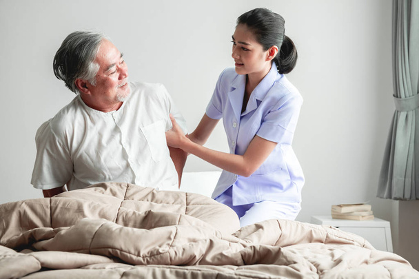 Senior man in bed trying to get up and nurse helping him. Old asian man and beautiful asian nurse woman in bedroom and open curtain. Senior home service concept. - Foto, Bild