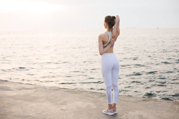 Kaunis tyttö urheilullinen ylä- ja valkoinen leggingsit kädestä takana merinäköalalla taustalla. Nuori nainen harjoittelee joogaa meren rannalla
 - Valokuva, kuva