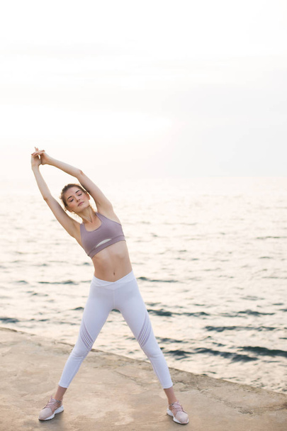 Nuori nainen urheilullinen ylhäältä ja valkoinen leggingsit tekee harjoituksia hämmästyttävä merinäköala taustalla. Huomaavainen tyttö venyttely meren rannalla
 - Valokuva, kuva