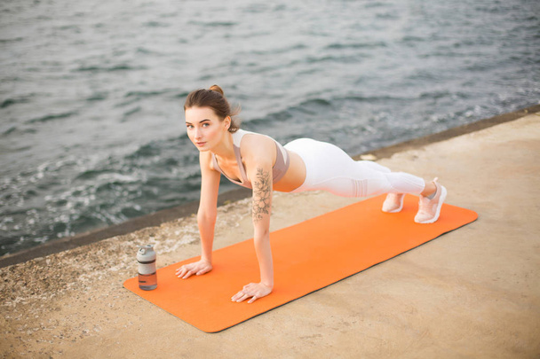 Jonge vrouw dromerig kijken in de camera praktizerende yoga aan zee. Mooi meisje in sportieve top en witte legging sport beoefening op oranje yoga mat met uitzicht op zee op de achtergrond - Foto, afbeelding