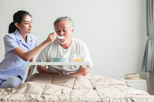 Young attractive nurse feeding breakfast to senior man in bed. Old asian male with white beard with young attractive asian woman caretaker. Senior home service concept. - Фото, изображение