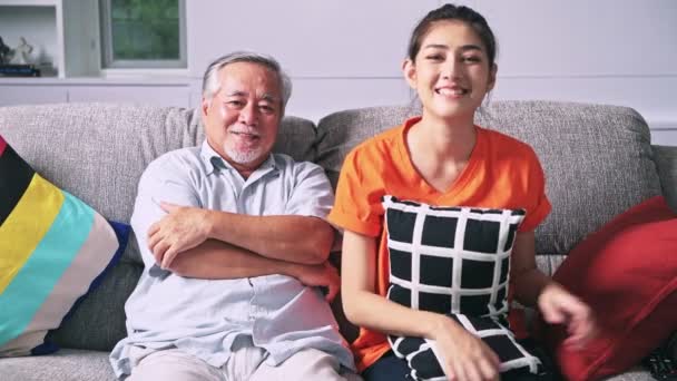 happy asian granddaughter with remote control and grandfather sitting on couch watching television - Materiaali, video