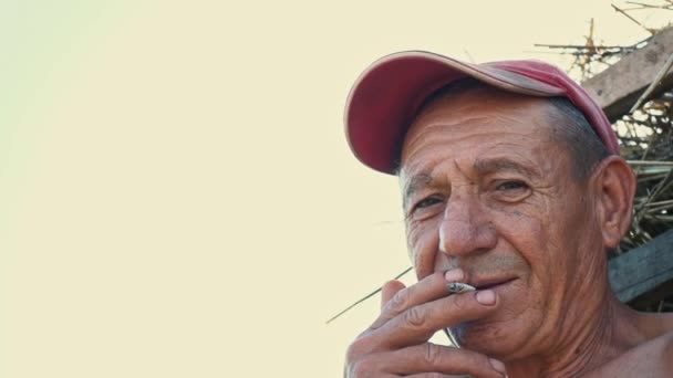 Retrato de um trabalhador fumante duro contra o pano de fundo de uma cena rural. Um agricultor com um boné fuma um cigarro
 - Filmagem, Vídeo