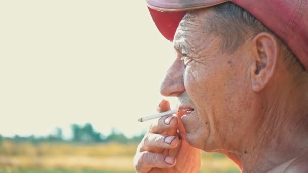 Πορτραίτο του καπνίσματος σκληρά εργαζόμενος φόντο την αγροτική σκηνή. Ένας αγρότης σε ένα καπάκι καπνίζει ένα τσιγάρο - Πλάνα, βίντεο