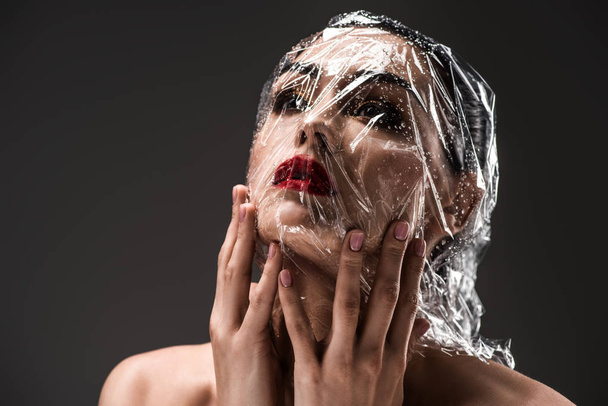 junge Frau mit in nasses transparentes Zellophan gehülltem Gesicht isoliert auf schwarz - Foto, Bild