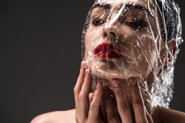 sensual joven con la cara envuelta en celofán transparente húmedo aislado en negro
  - Foto, Imagen