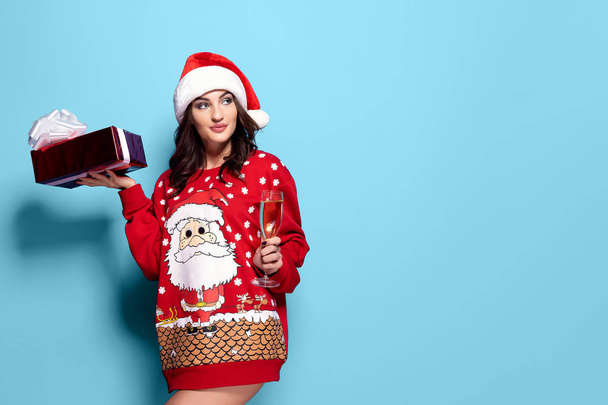 Pretty brunette woman in red pullover with Santa design, glass of champagne on blue background - Foto, immagini