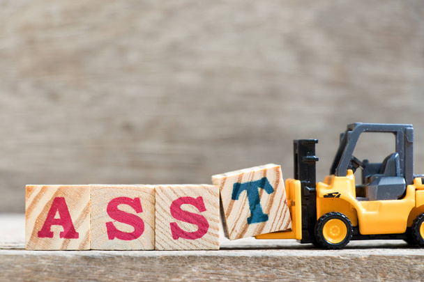 Spielzeuggabelstapler halten Buchstabenblock t in Wort asst (Abkürzung für Assistent) auf Holzgrund - Foto, Bild