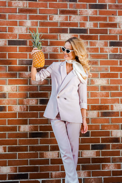 stylish young woman with pineapple standing in front of brick wall - Foto, imagen