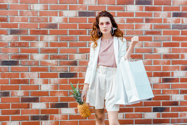 beautiful young woman with pineapple and shopping bags looking at camera in front of brick wall - 写真・画像