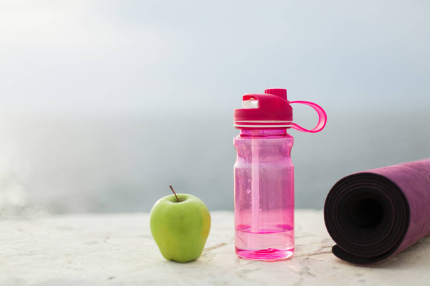Close-up van groene appel, roze sport fles en yoga mat over vage achtergrond - Foto, afbeelding