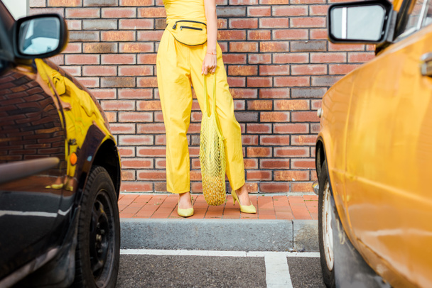 tiro recortado de mujer en ropa amarilla con piña en bolsa de hilo de pie en el aparcamiento delante de la pared de ladrillo
 - Foto, imagen