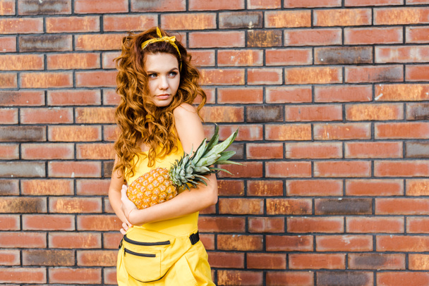attractive young woman in yellow clothes holding pineapple in front of brick wall and looking away - Фото, зображення