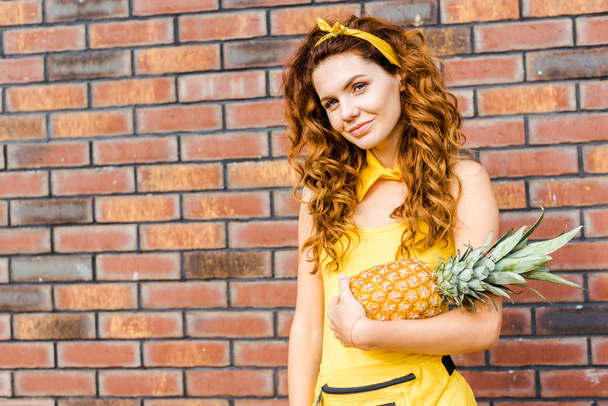 attractive young woman in yellow clothes holding pineapple in front of brick wall and looking at camera - Foto, Imagem