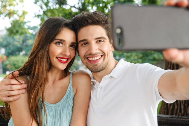 Fotografie mladých roztomilý milující pár sedí venku při make selfie pomocí mobilního telefonu. - Fotografie, Obrázek