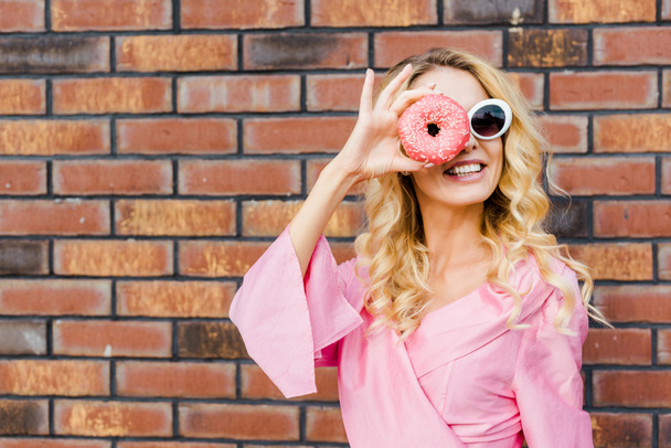 happy young woman in pink shirt looking at camera through donut in front of brick wall - Fotó, kép