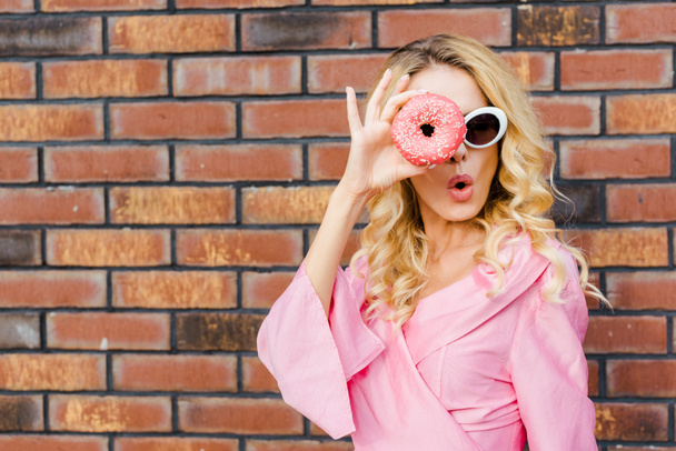 surprised young woman in pink shirt looking at camera through donut in front of brick wall - Photo, Image