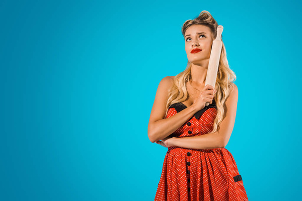 portrait of attractive pensive woman in pin up style dress with rolling pin looking away on blue background - Photo, Image