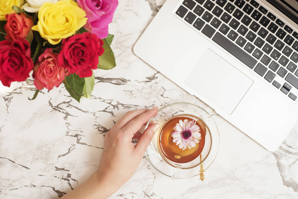 Feminine workplace concept. Freelance workspace in flat lay style with laptop, tea, flowers. Woman hand holding tea cup. Blogger working. Top view, bright, pink and gold. - Фото, изображение