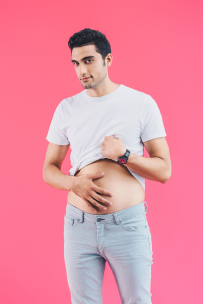 Veselý pohledný muž palming břicho a při pohledu na fotoaparát izolované na růžové - Fotografie, Obrázek