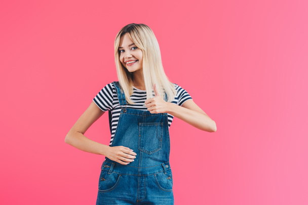 happy beautiful girl in denim overall showing thumb up and looking at camera isolated on pink - Foto, Bild