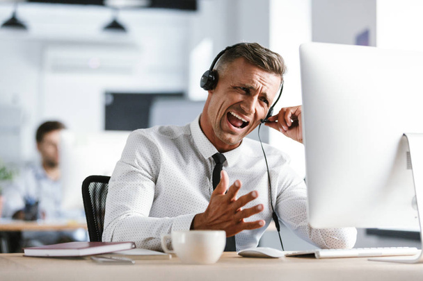 Photo of nervous businessman 30s wearing office clothes and headset yelling while sitting by computer - Valokuva, kuva