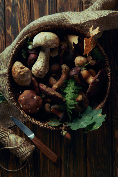 top view of fresh raw various edible mushrooms in basket, knife and rope on wooden table  - Fotografie, Obrázek