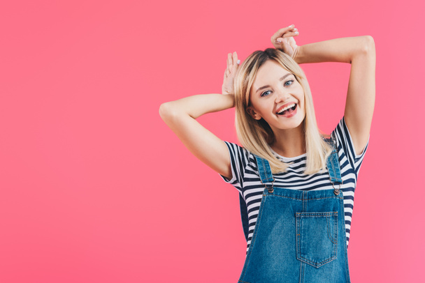 happy beautiful girl in denim overall showing bunny ears isolated on pink - Foto, Imagem