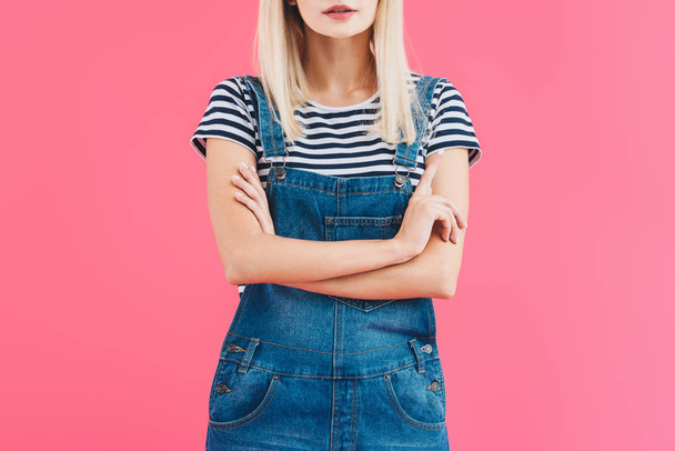 cropped image of girl in denim overall standing with crossed arms isolated on pink - Foto, Imagem