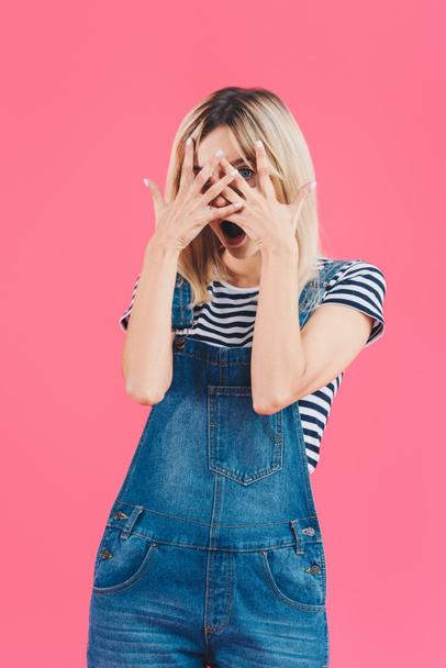 beautiful shocked girl in denim overall looking at camera through fingers isolated on pink - Fotoğraf, Görsel