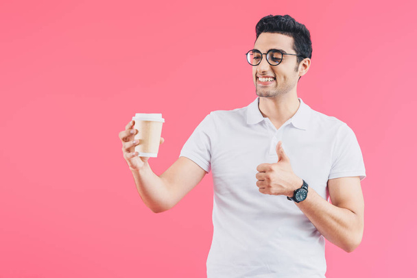 happy handsome man looking at coffee in paper cup and showing thumb up isolated on pink - Фото, изображение