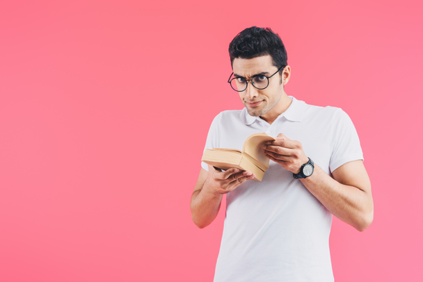 handsome skeptical student holding books and looking at camera isolated on pink - Foto, Imagen