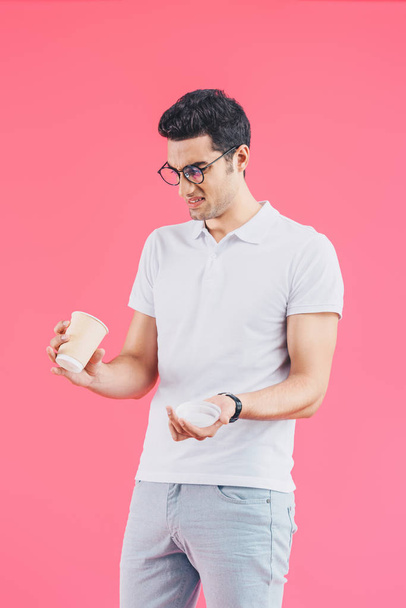 handsome grimacing man looking into disposable coffee cup isolated on pink - Zdjęcie, obraz