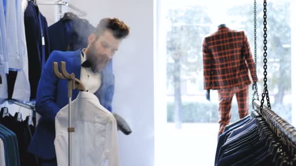 Elegante vendedor barbudo suaviza una camisa con vapor de ropa en la tienda de ropa masculina
. - Imágenes, Vídeo