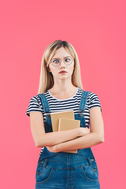 portrait of grimace young student in eyeglasses with books isolated on pink - 写真・画像