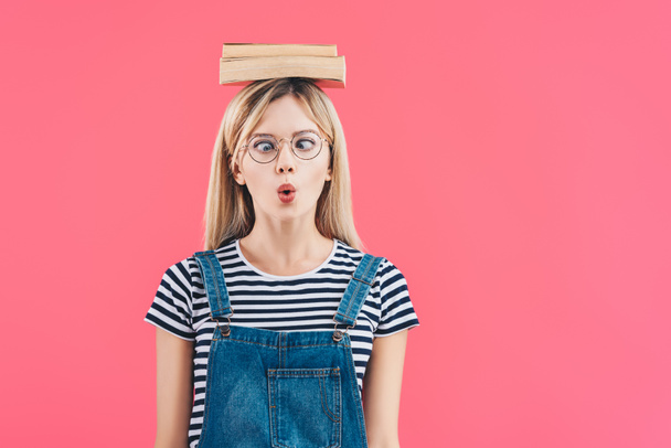 portrait of young grimace student in eyeglasses with books on head isolated on pink - 写真・画像