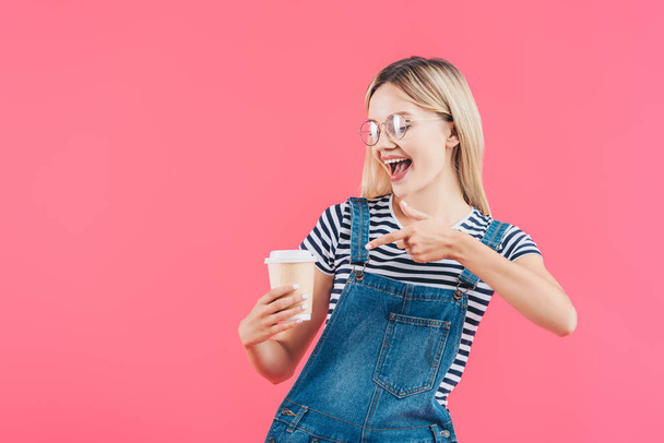 portrait of young woman pointing at coffee to go in hand isolated on pink - Photo, Image