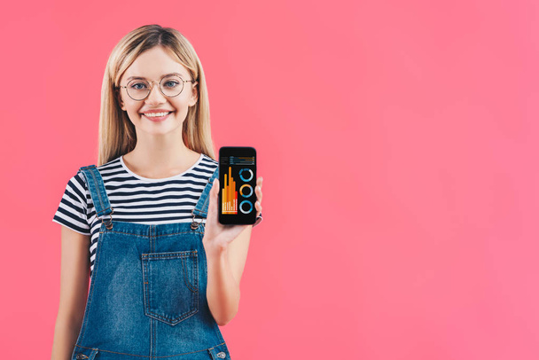portrait of smiling woman in eyeglasses showing smartphone isolated on pink - 写真・画像