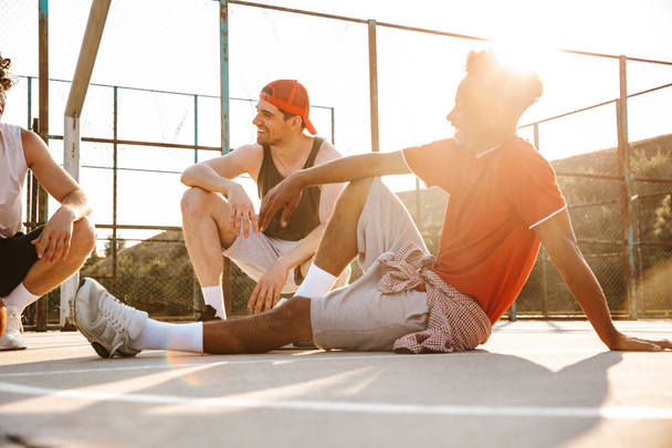 Group of young happy multiethnic men basketball players resting at the sport ground, talking - 写真・画像