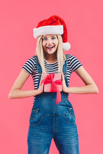 portrait of smiling woman in santa claus hat holding gift isolated on pink - Фото, зображення